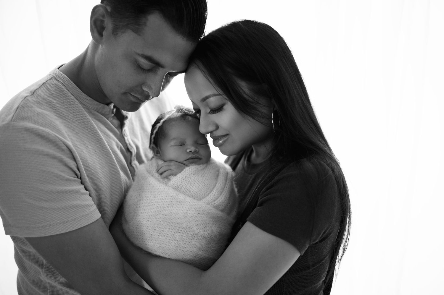 Black and white portrait of parents tenderly holding their newborn baby swaddled in white.