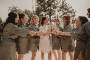 Wedding party in gray robes toasting champagne glasses together outdoors with pine trees in background.