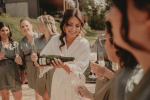 A group celebrates with champagne outdoors as someone in white pours drinks during pre-wedding festivities.