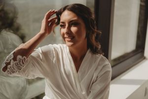 Person in white lace robe smiles while getting ready by window in morning light.