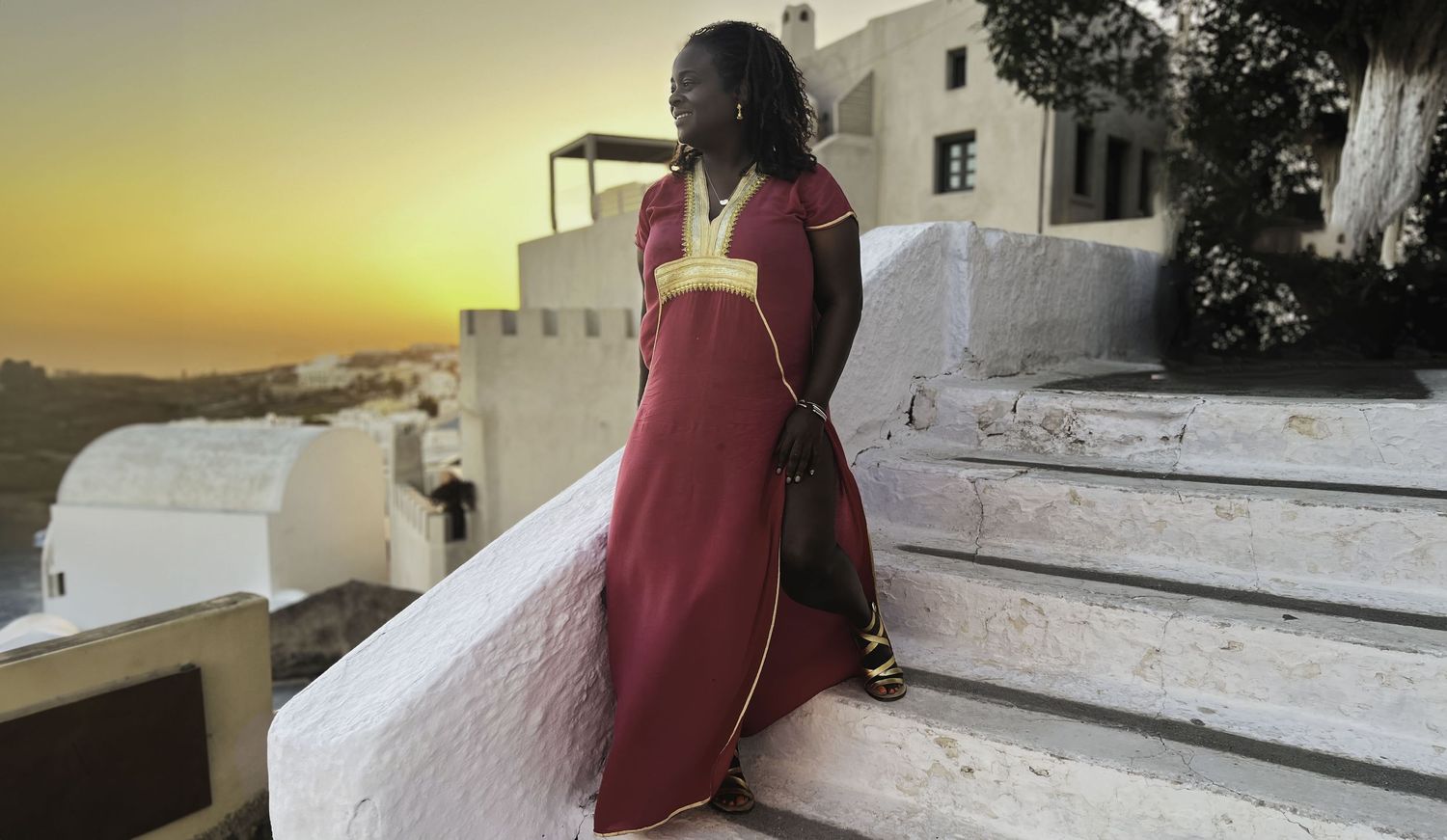 Tall woman in a red and gold traditional dress standing on white steps at sunset.