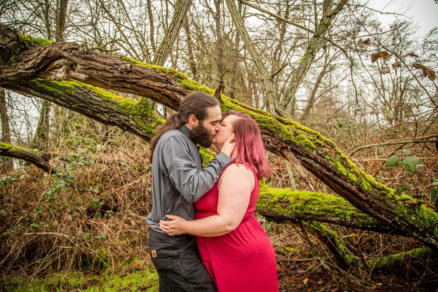 A couple embraces in front of a moss-covered fallen tree in a moody forest setting.