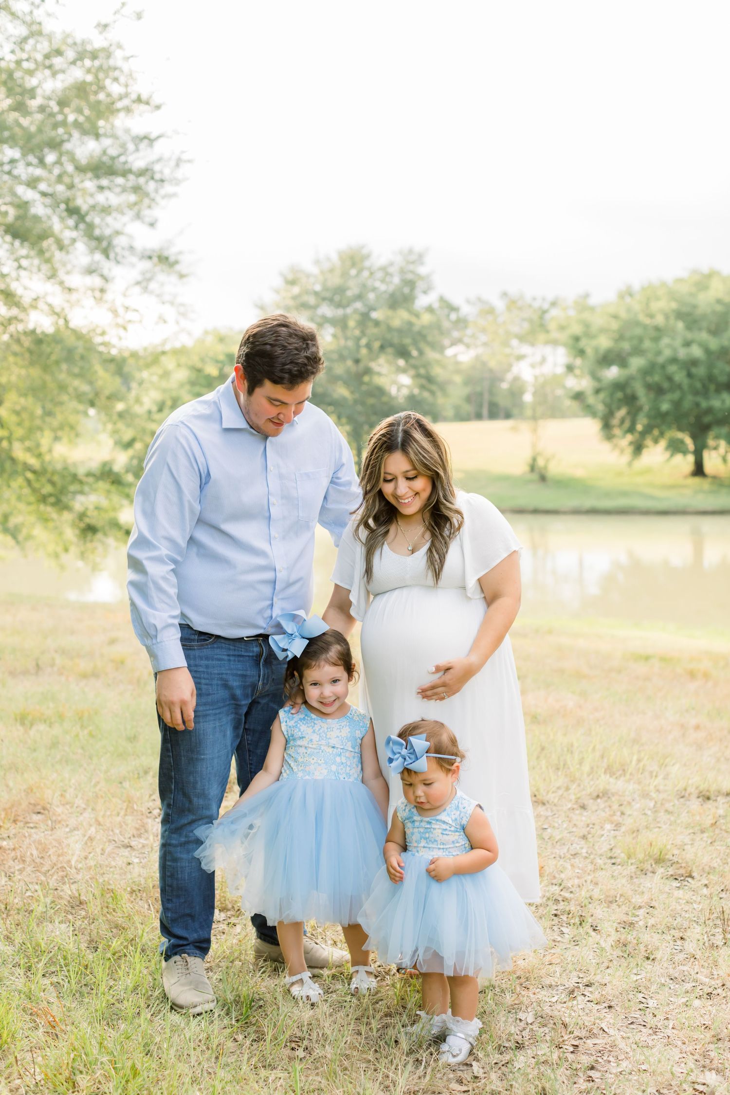 Family enjoying a sunny day by a pond with two children in matching blue dresses and a mother in a white dress.