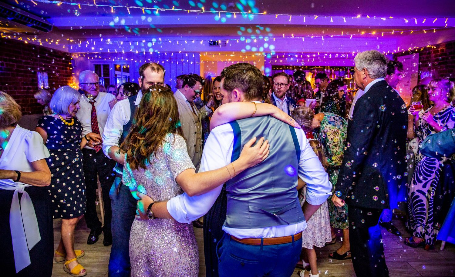 Couples slow dance under twinkling lights at an evening wedding reception.