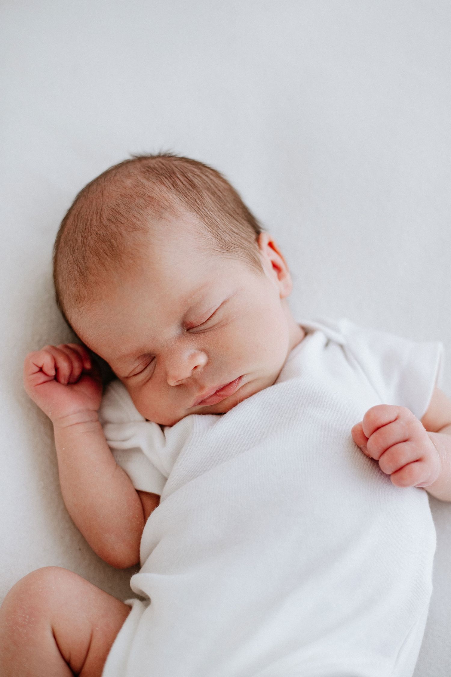 Sleeping baby in white onesie lying on light background with hand near face.