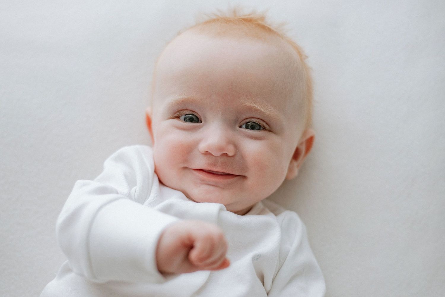 Happy baby in white outfit smiling at camera against light background.
