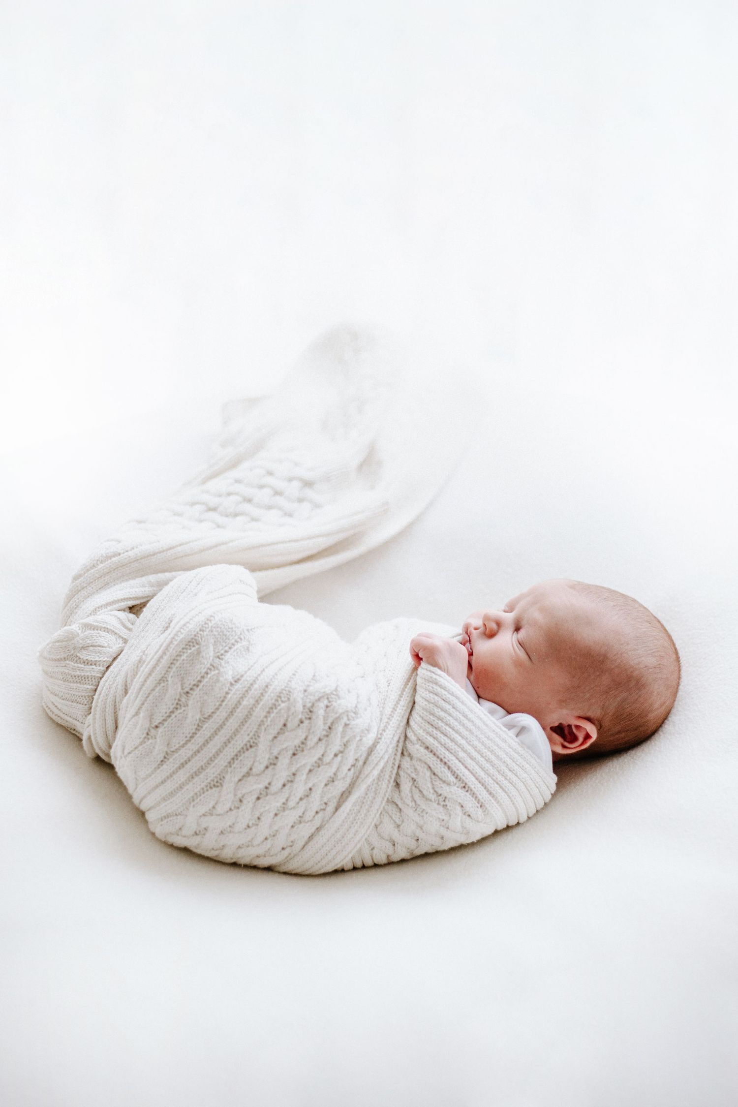 Sleeping newborn wrapped in white cable knit blanket on white background.