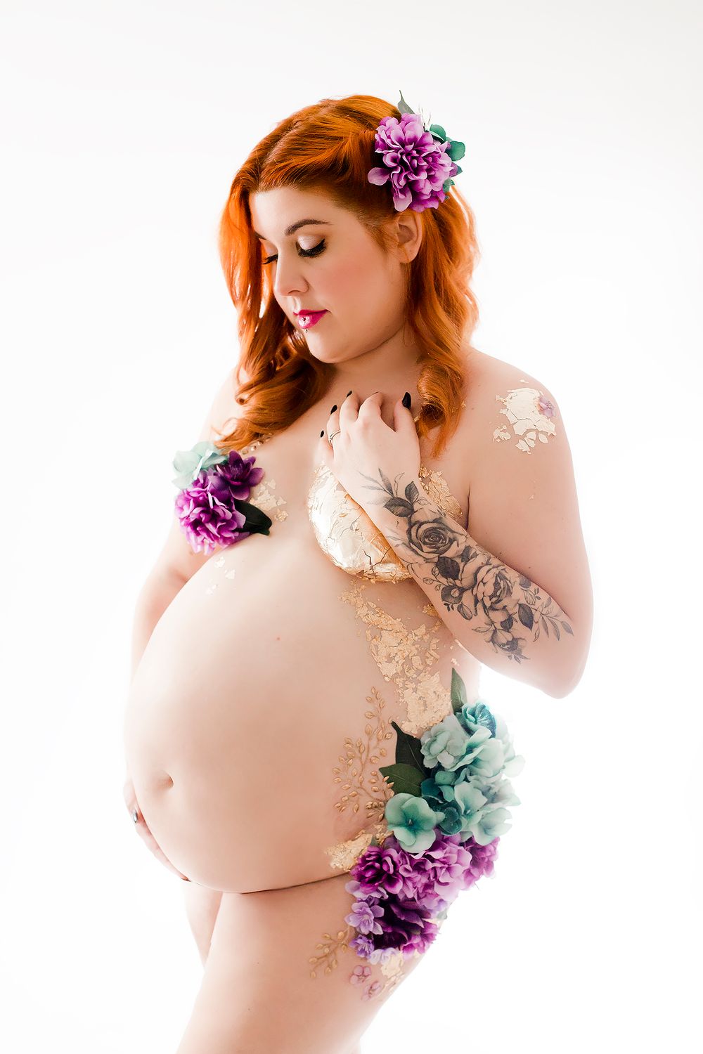 Artistic maternity photo featuring purple flowers, decorative body paint, and soft lighting against white background.