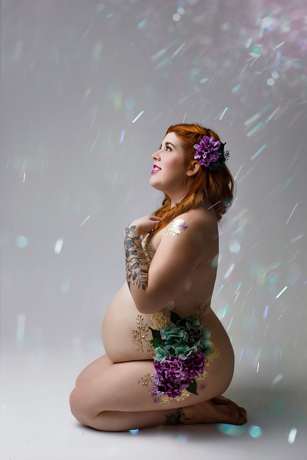 Artistic maternity photo with purple flowers and sparkling light effects against a gray backdrop.