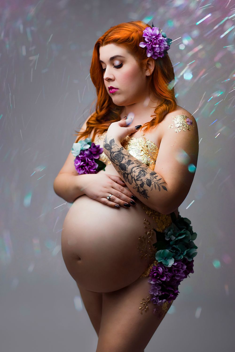 Artistic maternity photo featuring purple flowers, body art, and ethereal lighting against a gray backdrop.