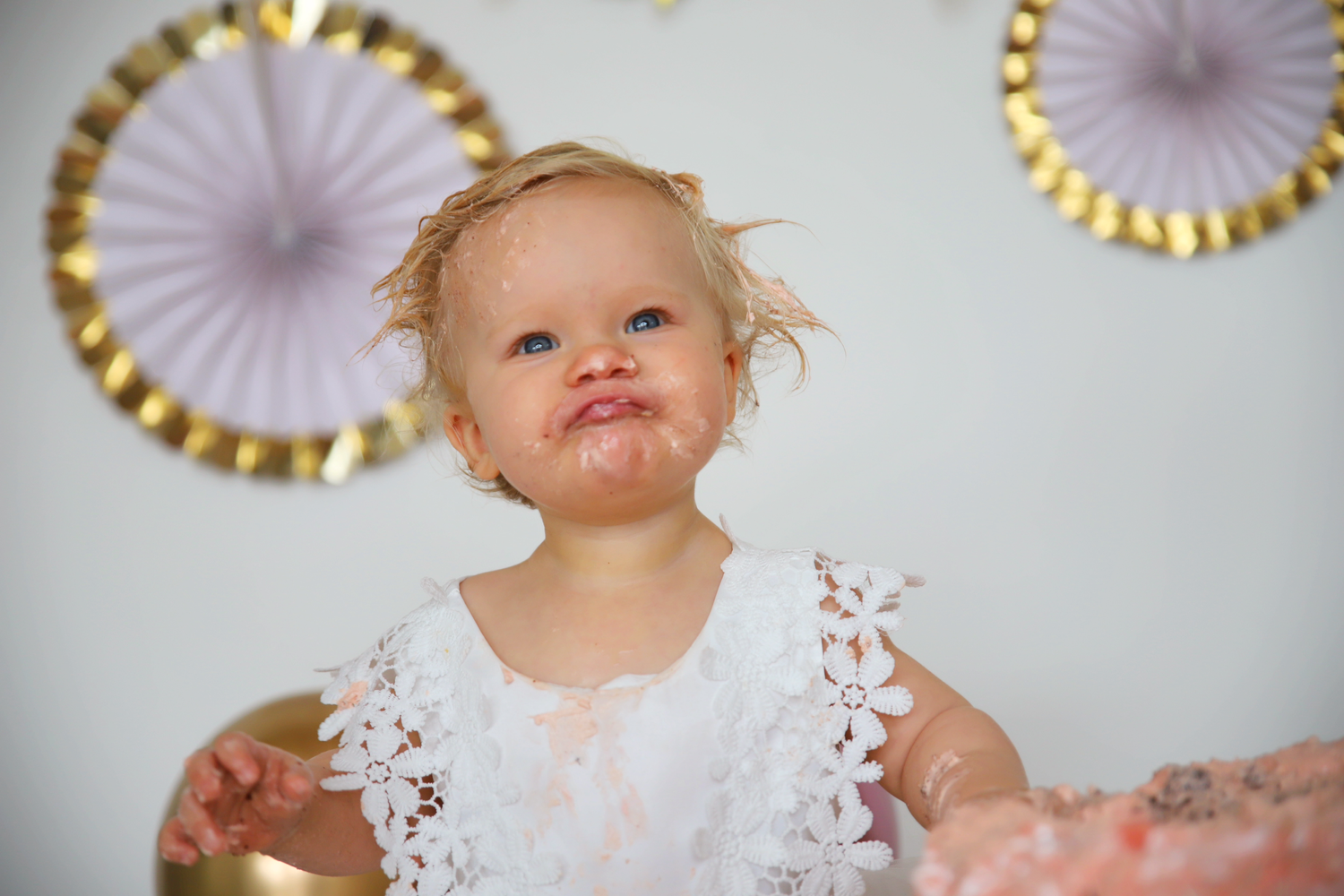 Baby in white lace dress with pink and gold party decorations in background.