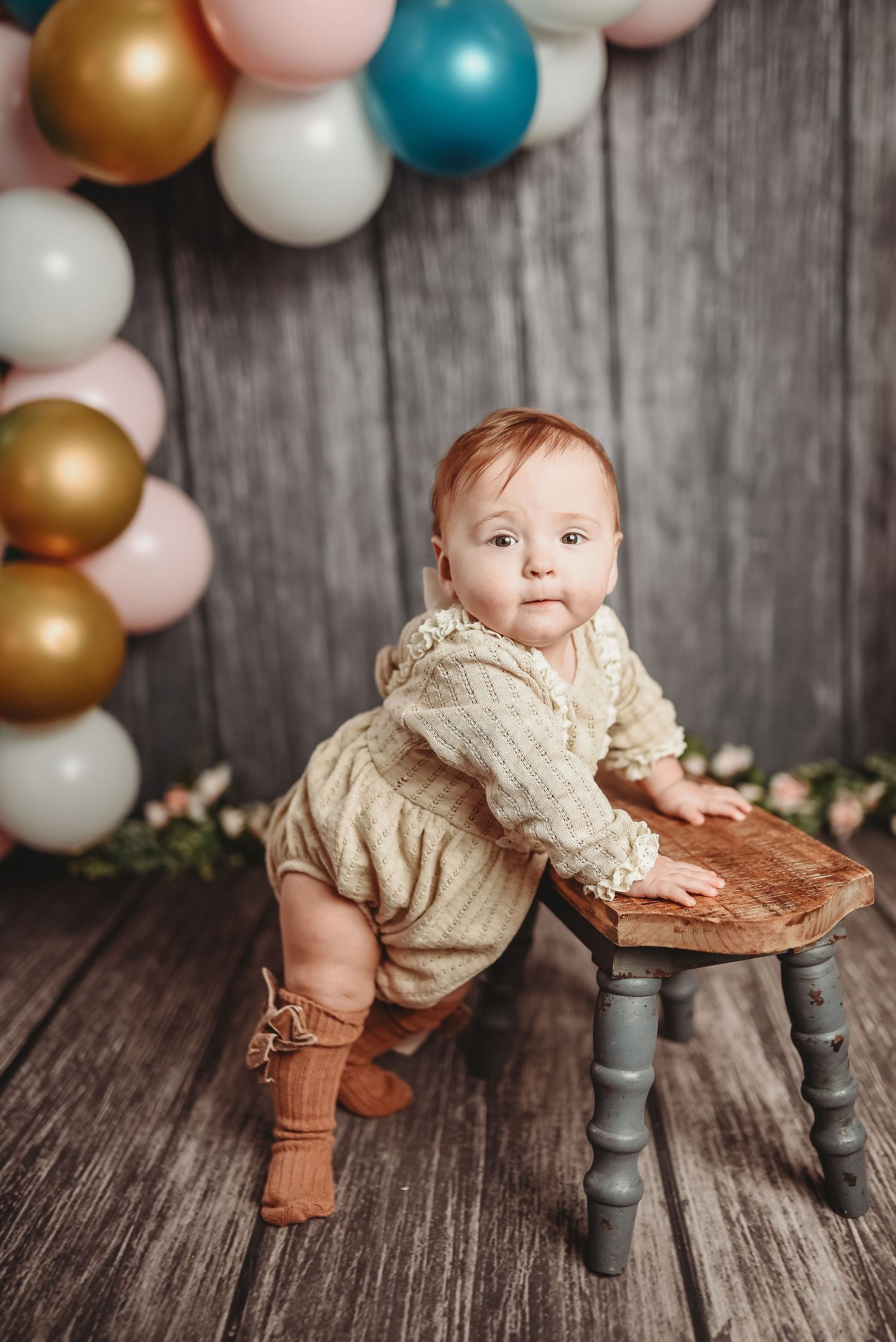 Baby in cream outfit stands by gray stool with balloon arch backdrop in pink, gold and blue.