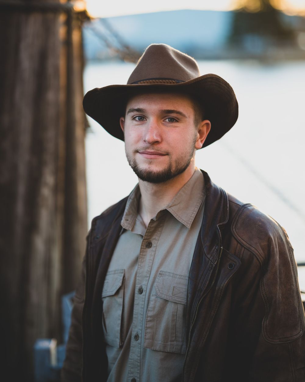 Young man in a brown cowboy hat and leather jacket posing near a waterfront.