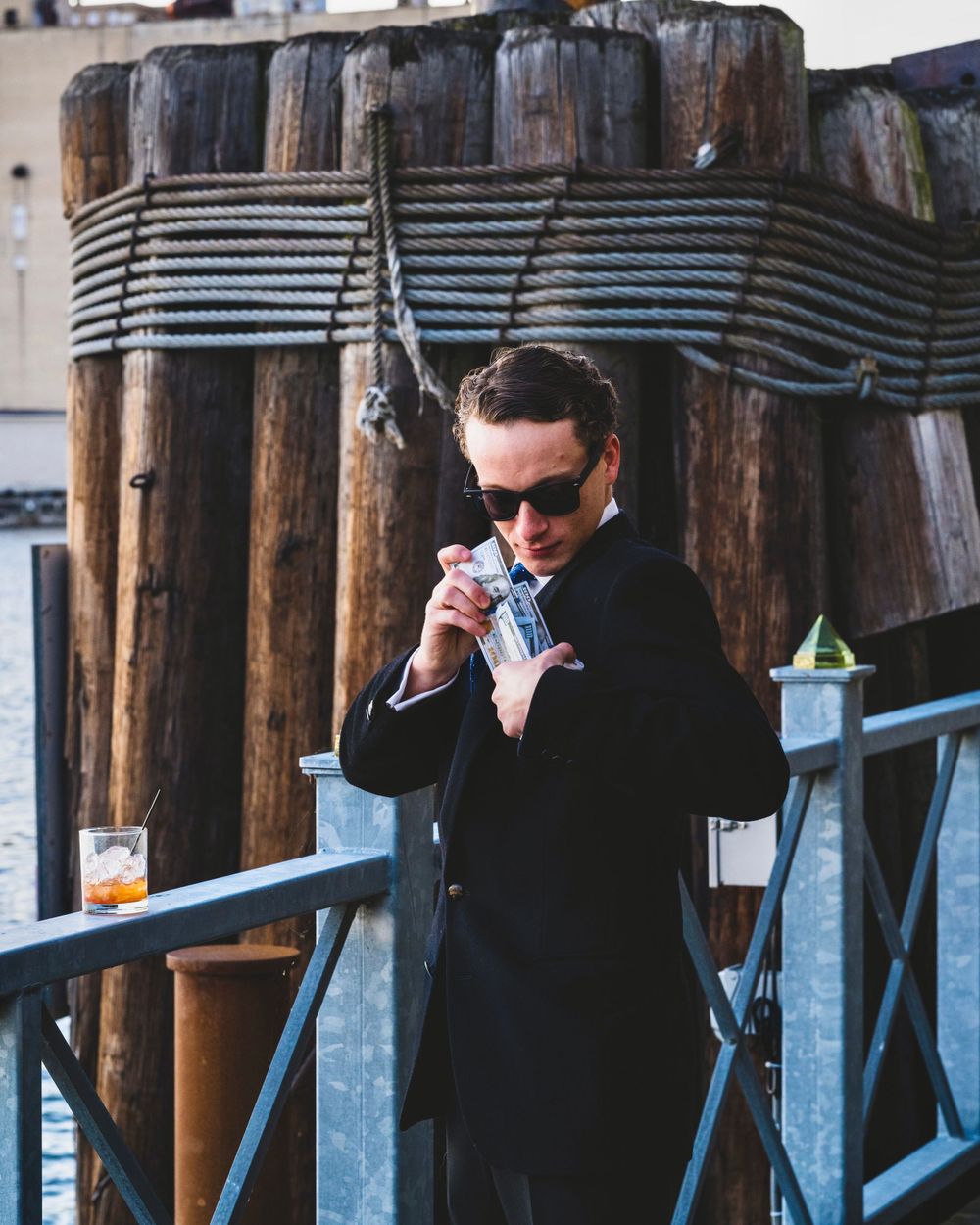 Man in black coat and sunglasses opening a packet near a wooden pier structure.