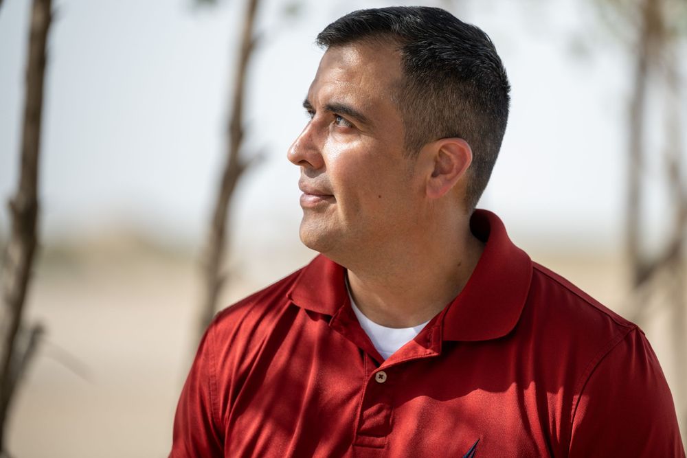 Hispanic man in red polo shirt gazing thoughtfully into distance outdoors.