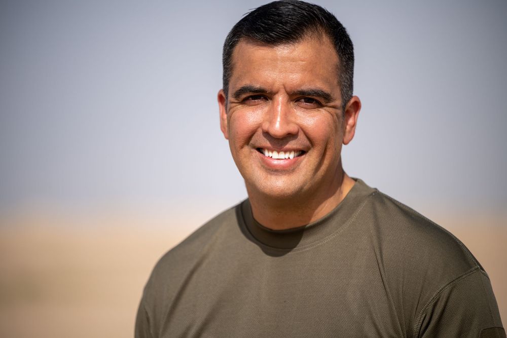 Smiling Hispanic man in olive t-shirt with sandy desert background outdoors.