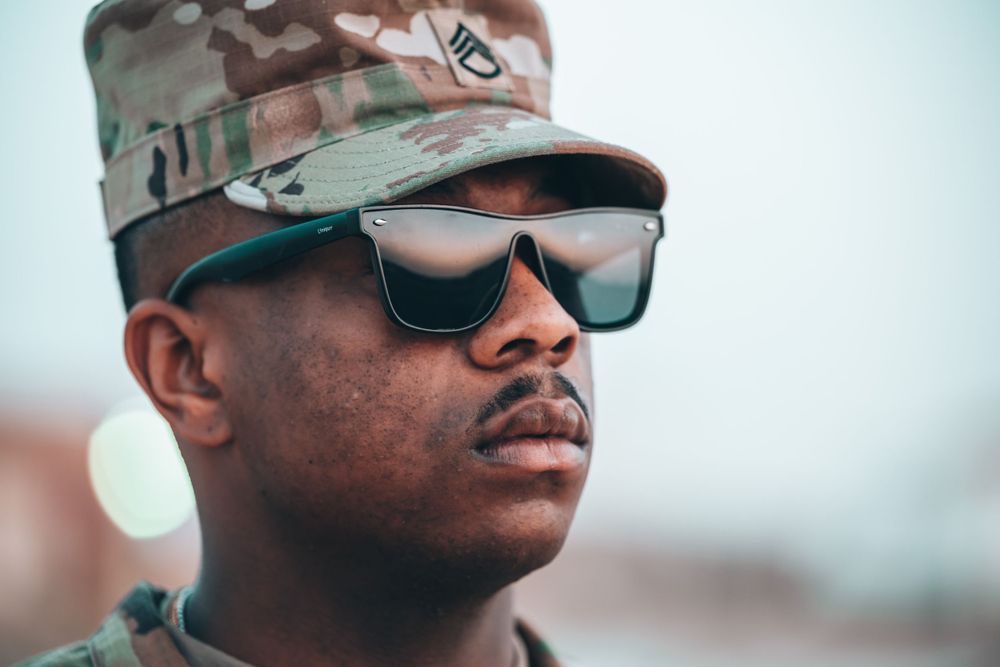 Close-up portrait of US Army soldier in camouflage cap wearing reflective sunglasses outdoors.