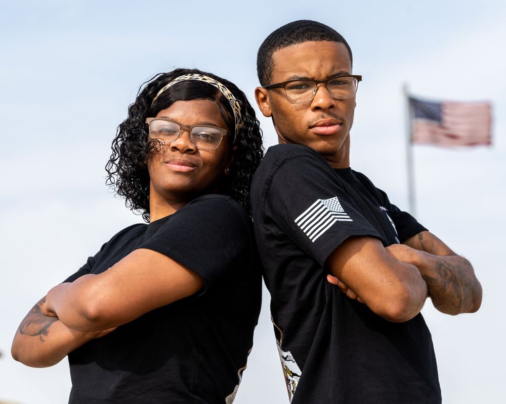 Two young Black Americans in black shirts standing back-to-back with US flag in background.