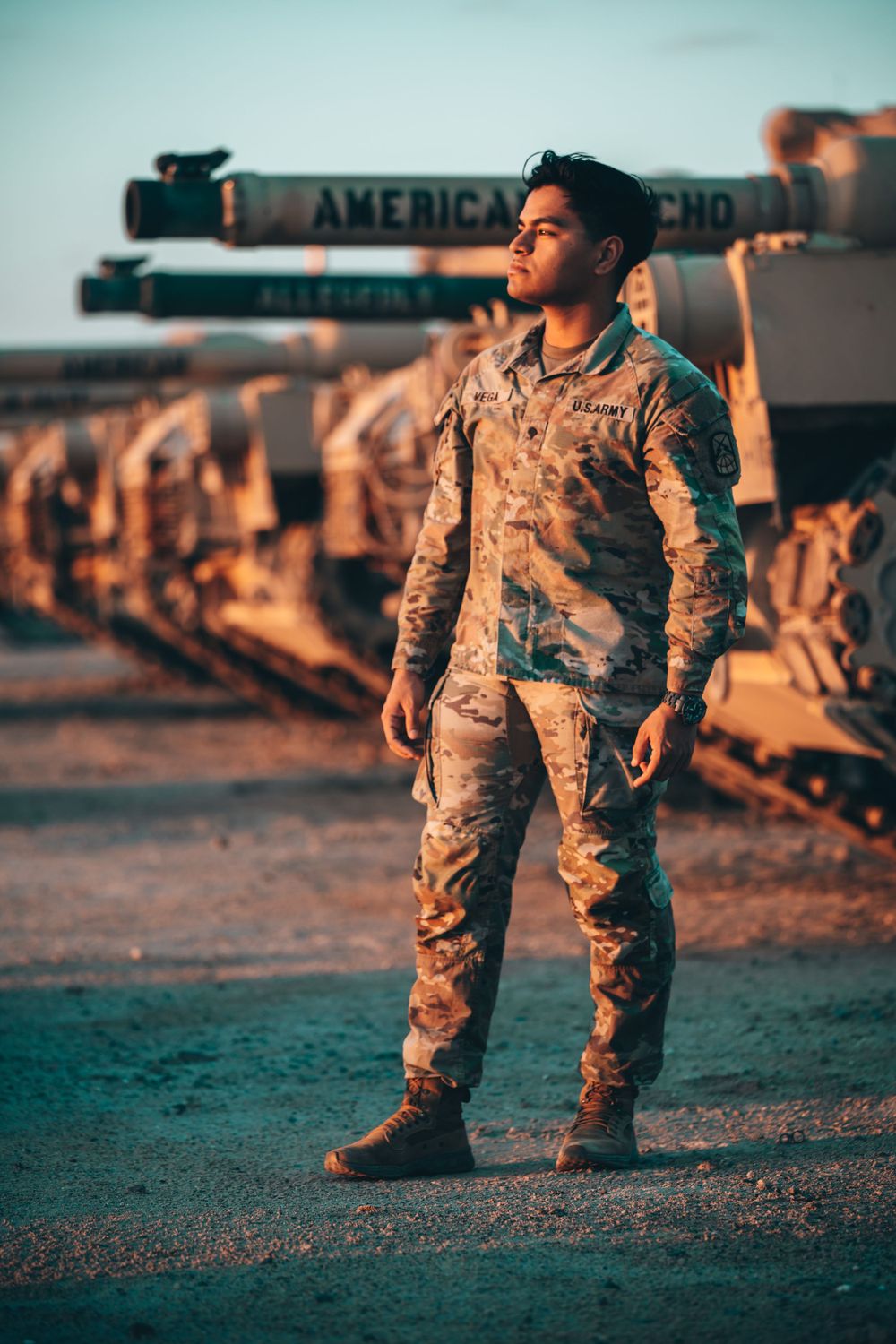 US Army soldier in OCP uniform standing before military tanks at golden hour.