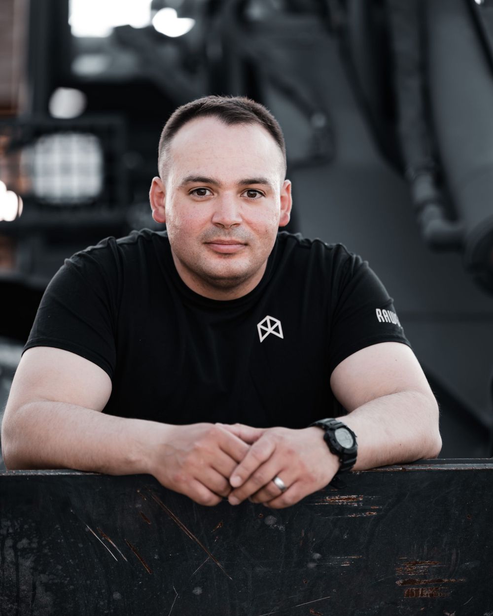 Man in black t-shirt leaning on military vehicle with hands clasped, smiling at camera.