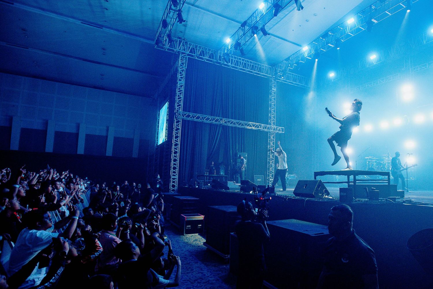 Concert performers silhouetted against blue stage lights with crowd in foreground.