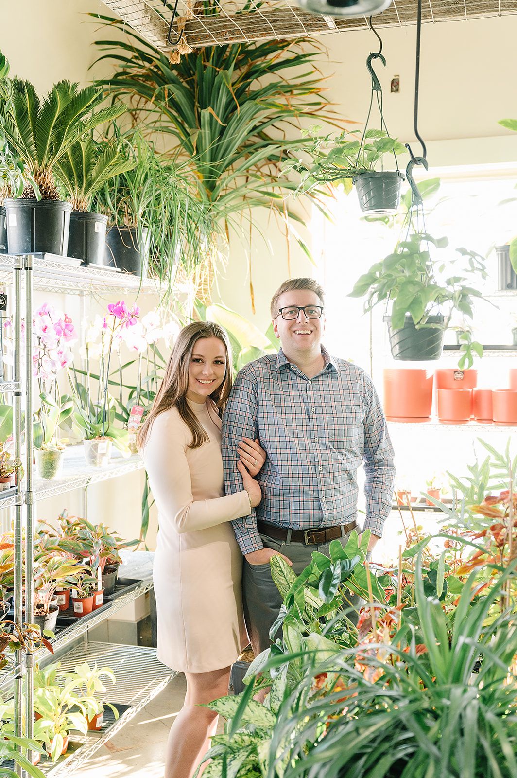 Couple posing together in a greenhouse surrounded by tropical plants and hanging orchids.