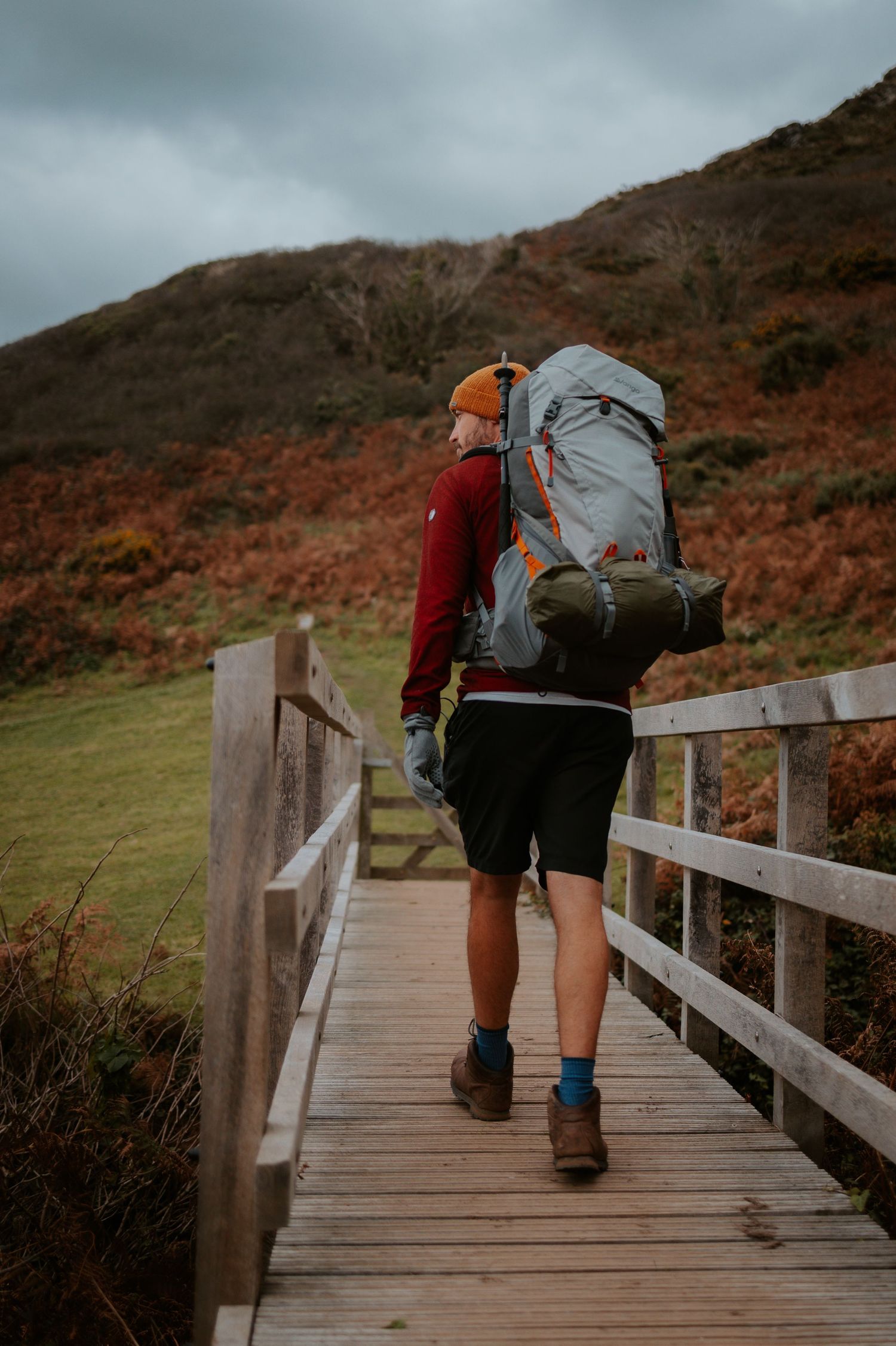Hiker with large backpack walks along wooden boardwalk through coastal landscape.