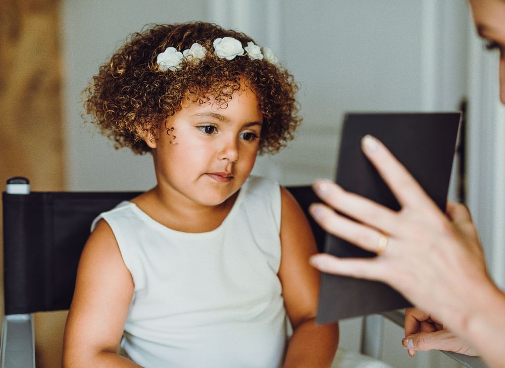 Child with curly hair and white top looking at someone holding a device in an indoor setting.