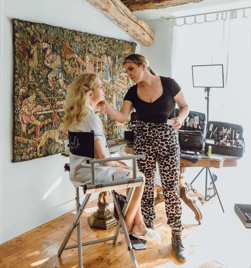 Two people doing makeup preparation in room with decorative tapestry.