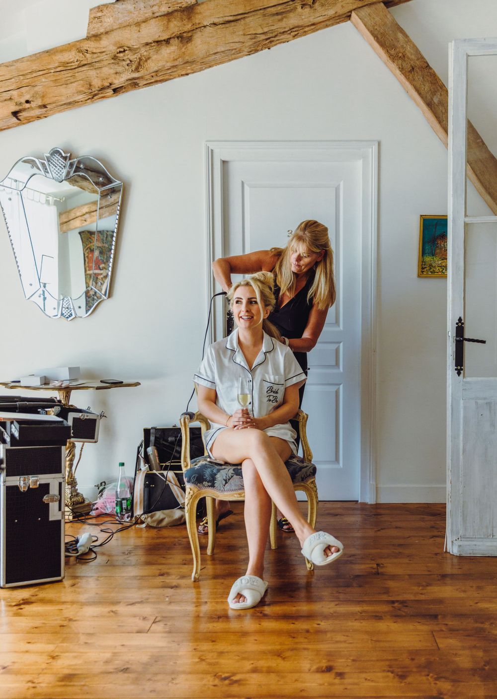Wedding preparation scene with hair styling in a rustic room with wooden beams and hardwood floors.