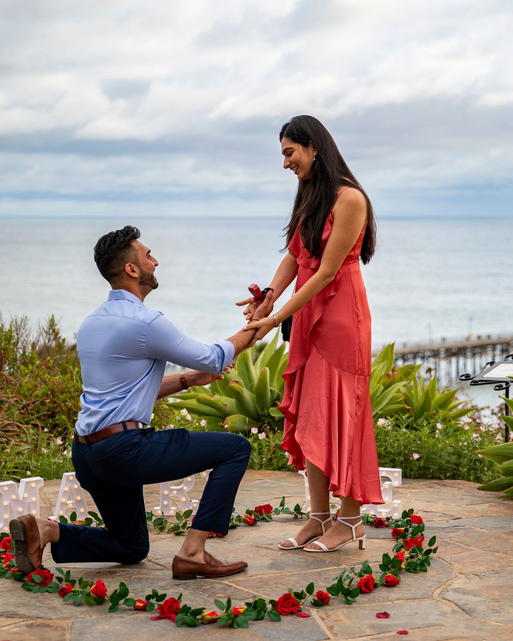 Orange County photographer captures Man proposing to woman with rose petal heart on scenic ocean overlook patio.