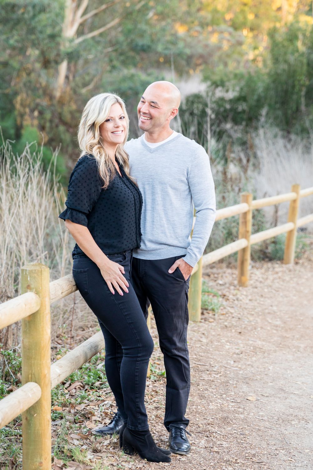 Orange County photographer captures Smiling couple stands on scenic nature path by wooden fence, woman in black outfit, man in grey sweater.