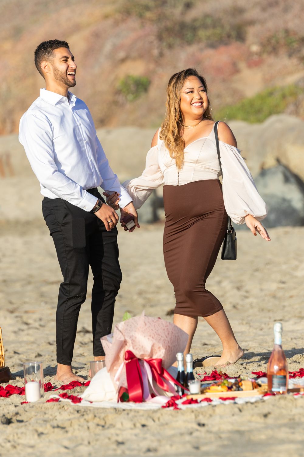 Orange County photographer captures Couple walking hand in hand on a beach with a romantic picnic setup nearby.