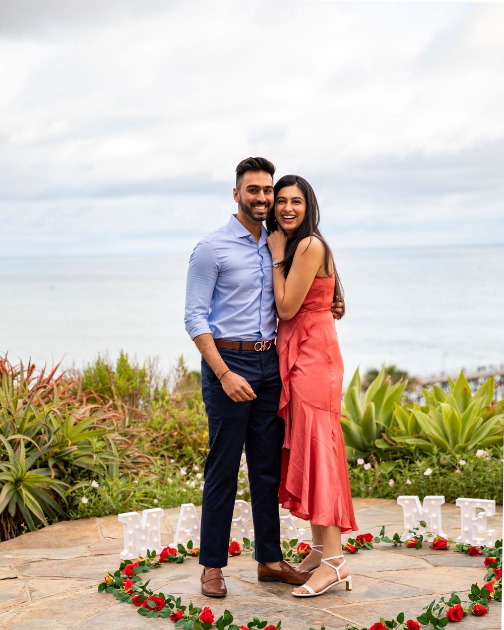 Orange County photographer captures Happy engaged couple posing with rose petal heart arrangement by ocean view.