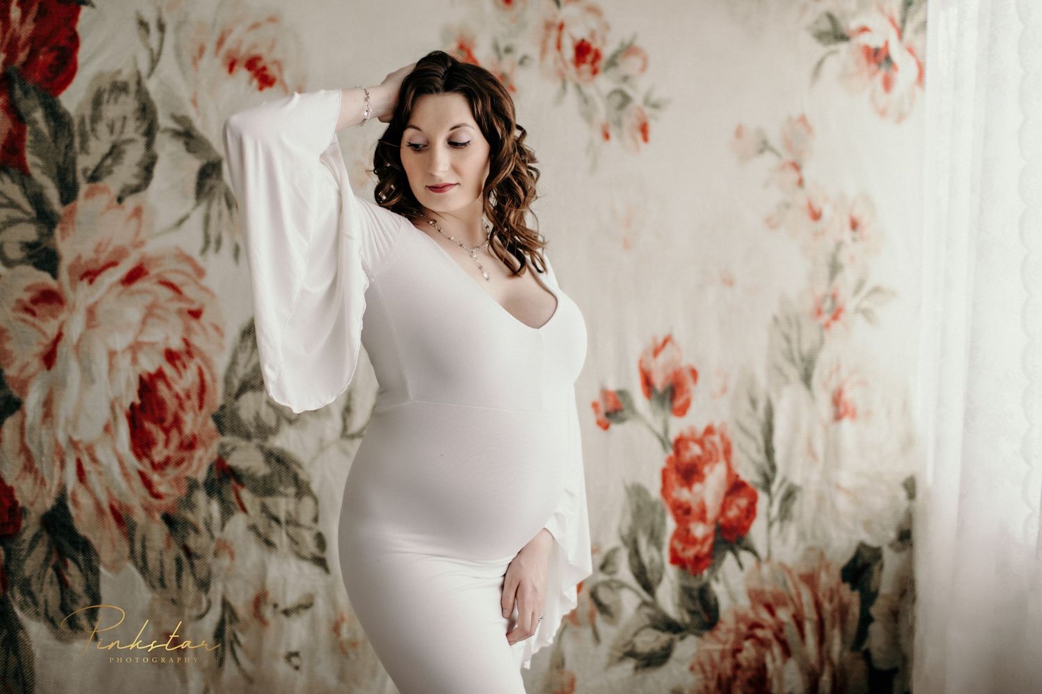 A maternity portrait featuring a white long-sleeve dress against a floral wallpaper background.