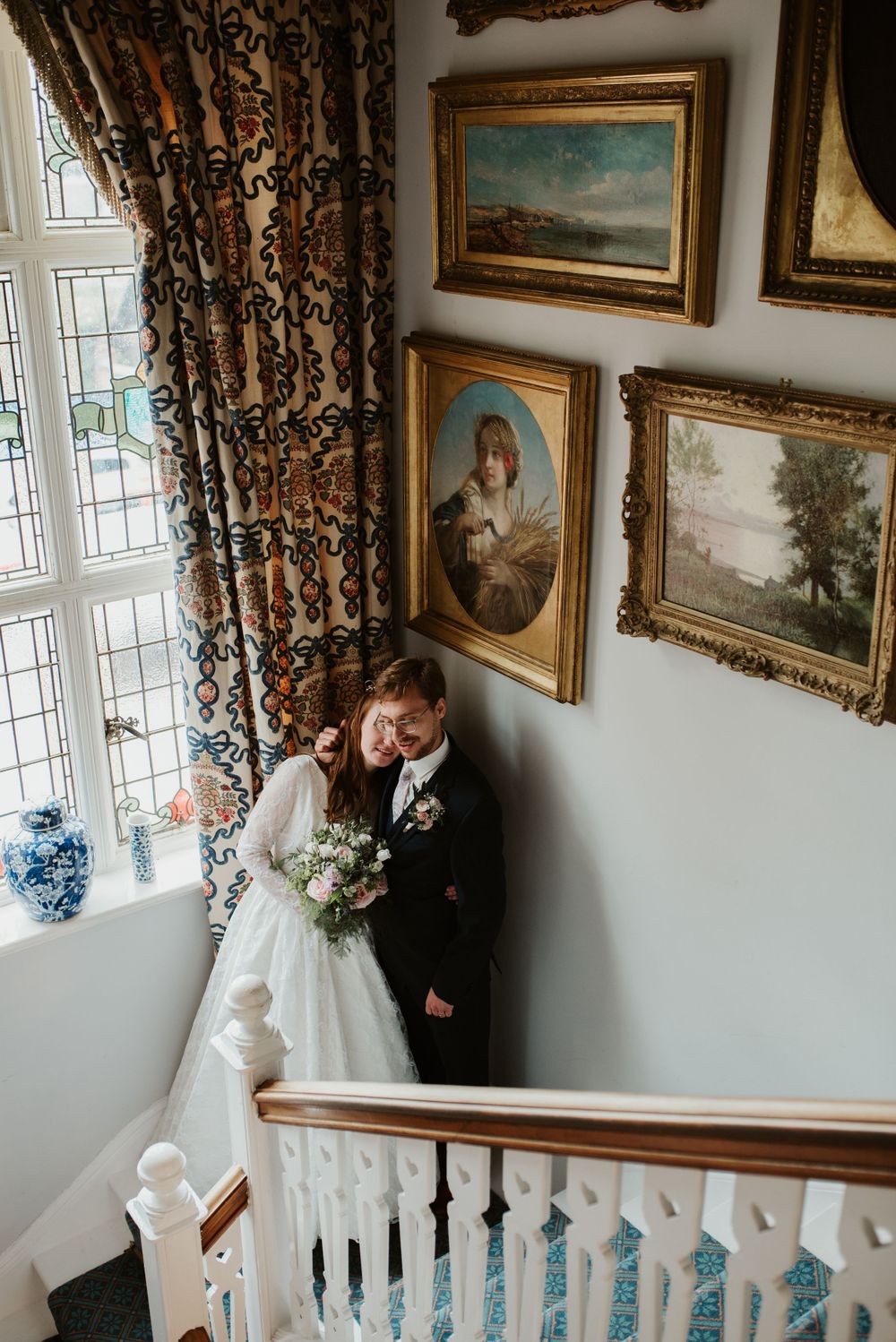Wedding couple poses in an elegant stairwell with ornate framed artwork and curtains.