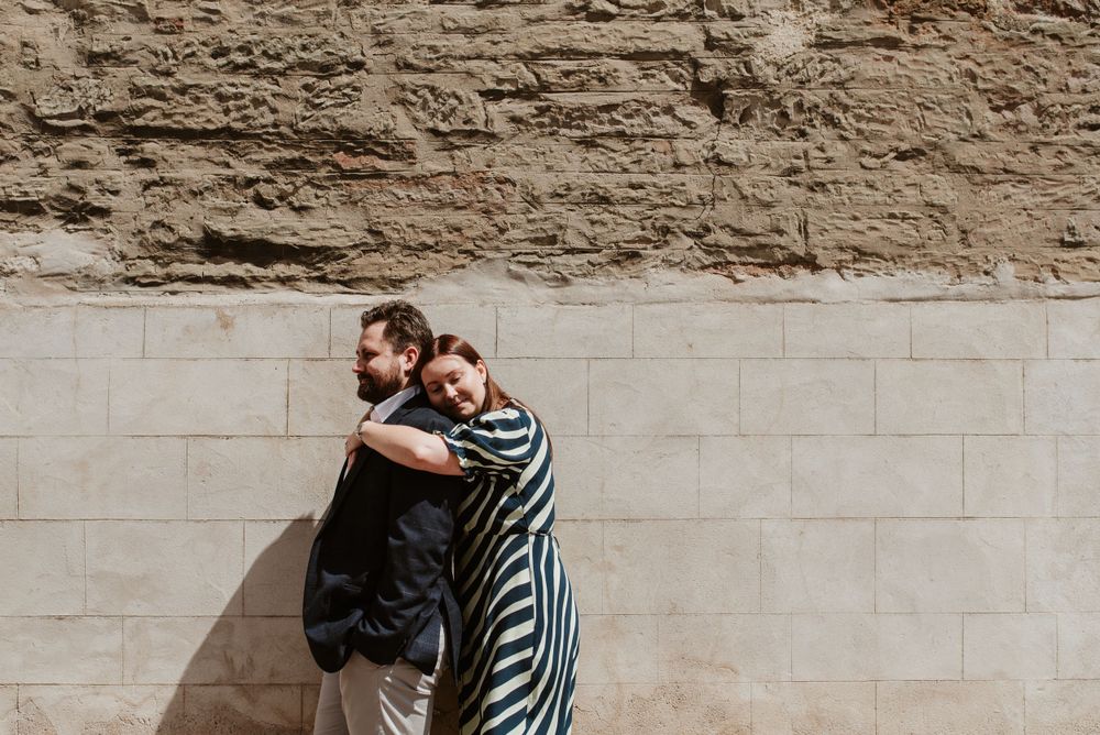 Couple embracing against stone wall in striped dress and dark clothing.