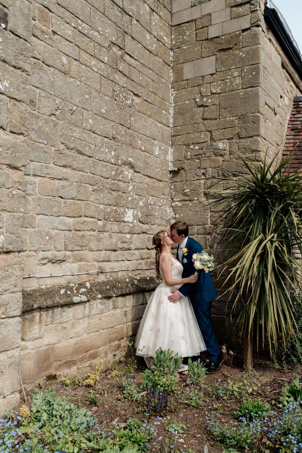 A romantic wedding photo of a couple embracing against a stone wall with tropical plants.