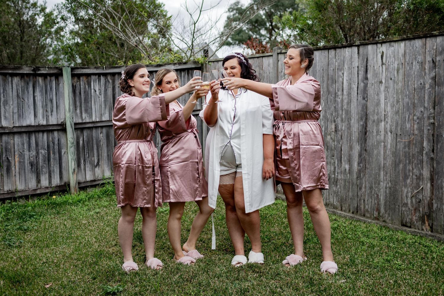 Wedding party members in pink robes raise glasses for a celebratory toast outdoors.