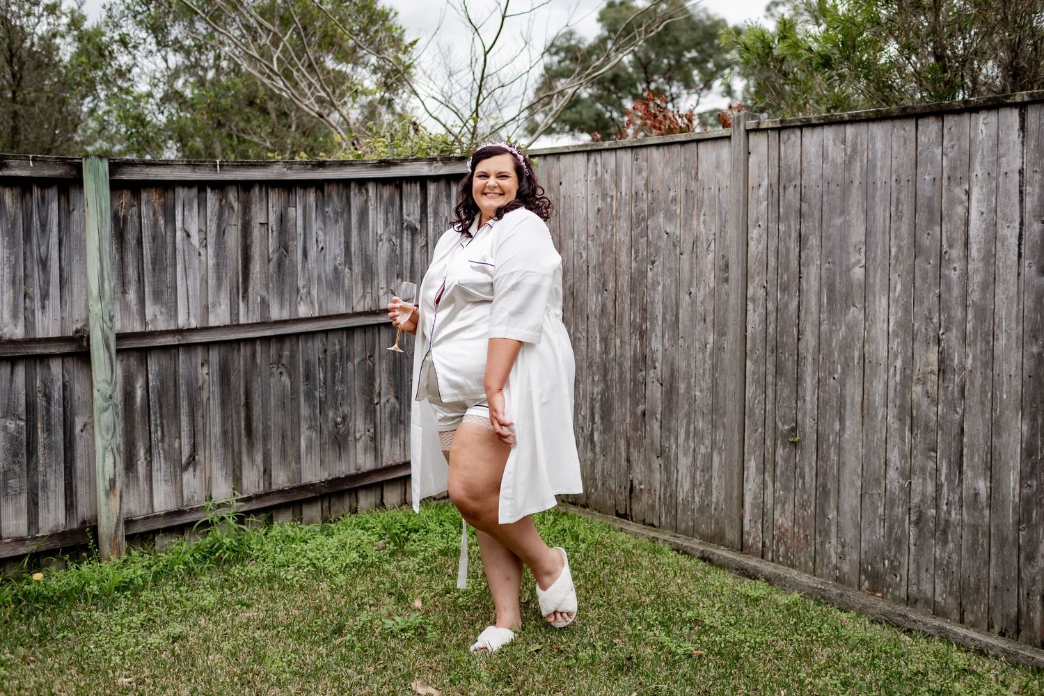 A person in a white robe poses in a backyard with a wooden fence backdrop.