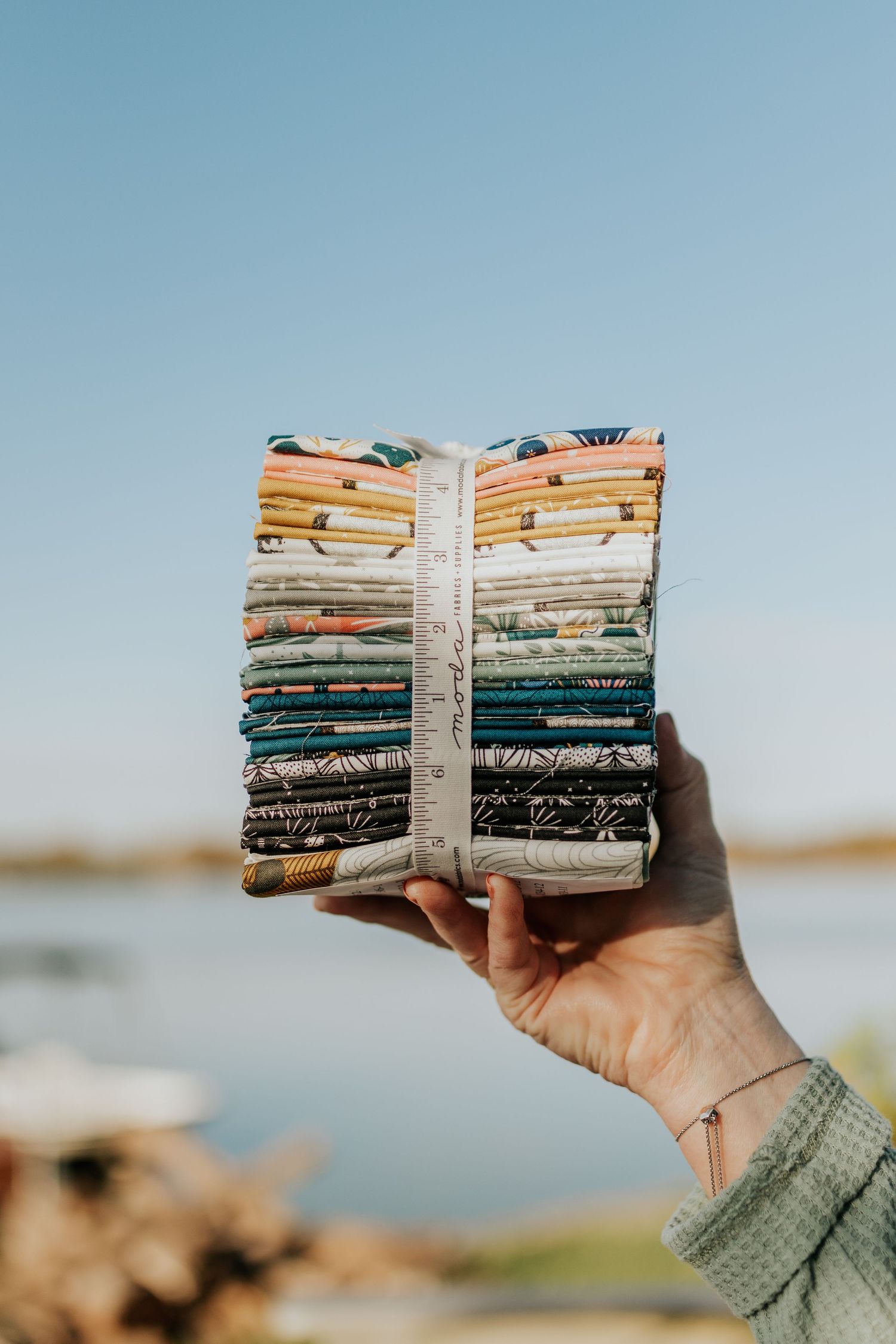 A longarm quilter holds up a stack of fat quarters in front of a blue sky during a personal branding photoshoot in KC.