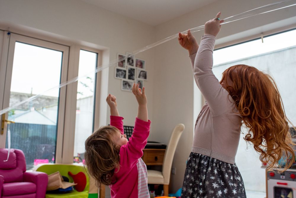Two people playing with a string or rope by large windows in a bright room.