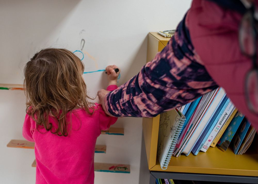 Child in pink shirt writing or drawing on wall mounted paper.