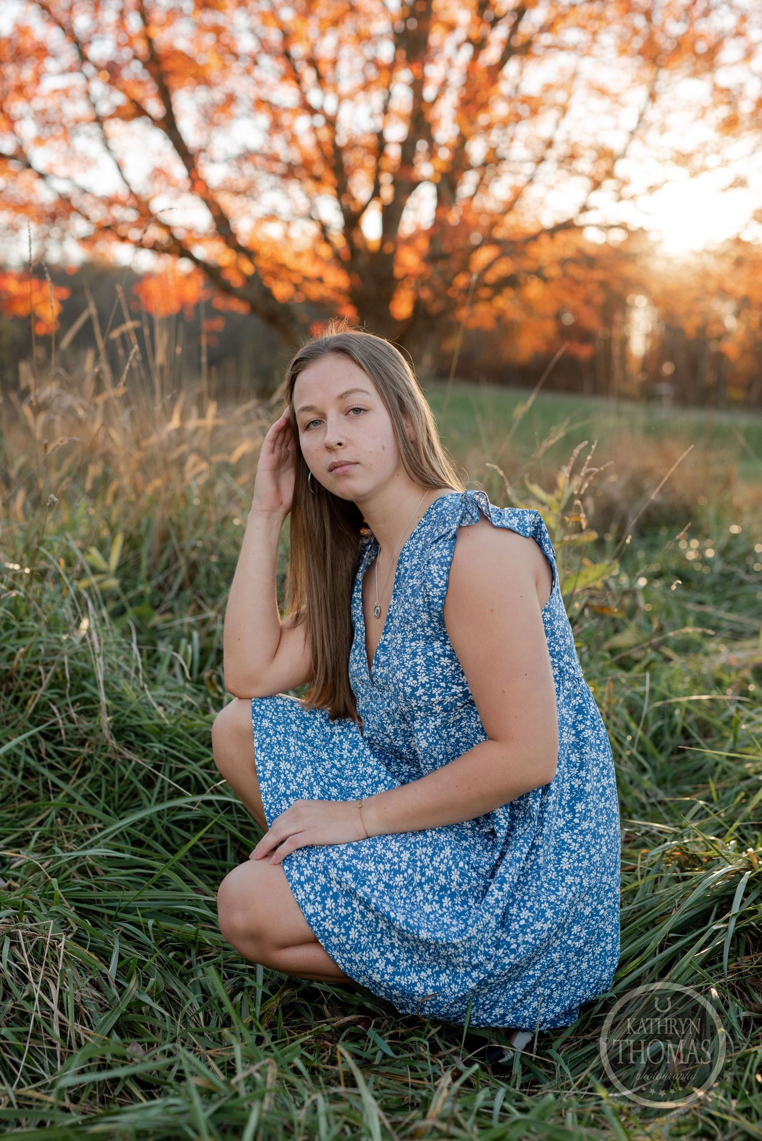 A blue floral dress in tall grass with autumn trees glowing at sunset.