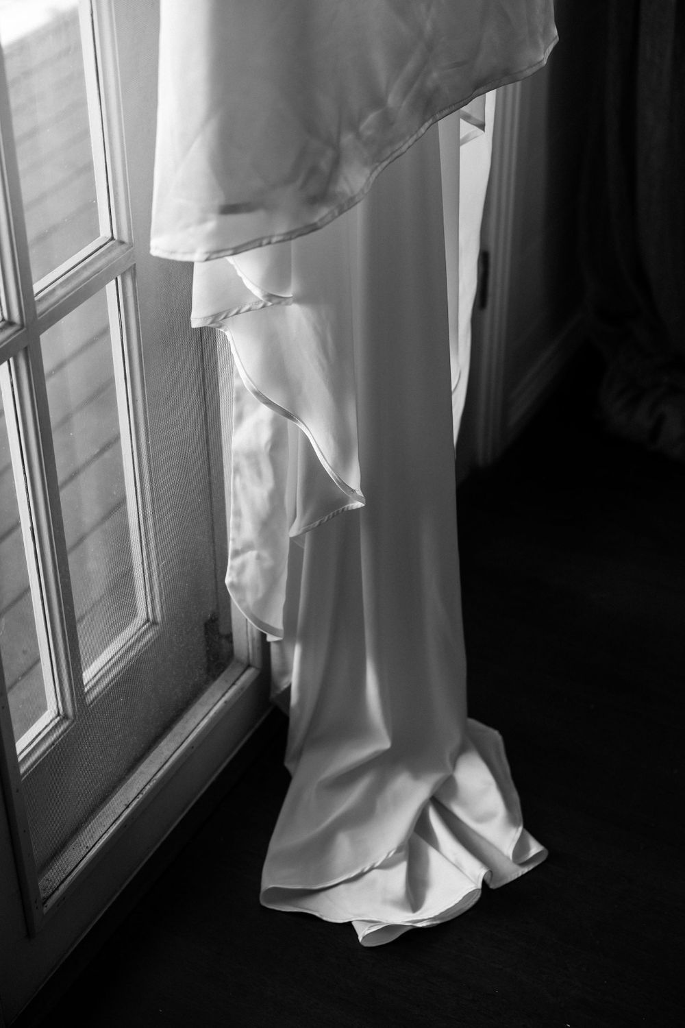Detail shot of a white wedding dress train flowing against a window with dramatic lighting.