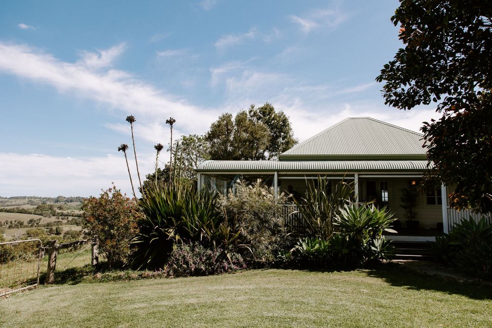 A rustic countryside house with a wraparound porch and tropical plants sits on a grassy hill under blue skies.