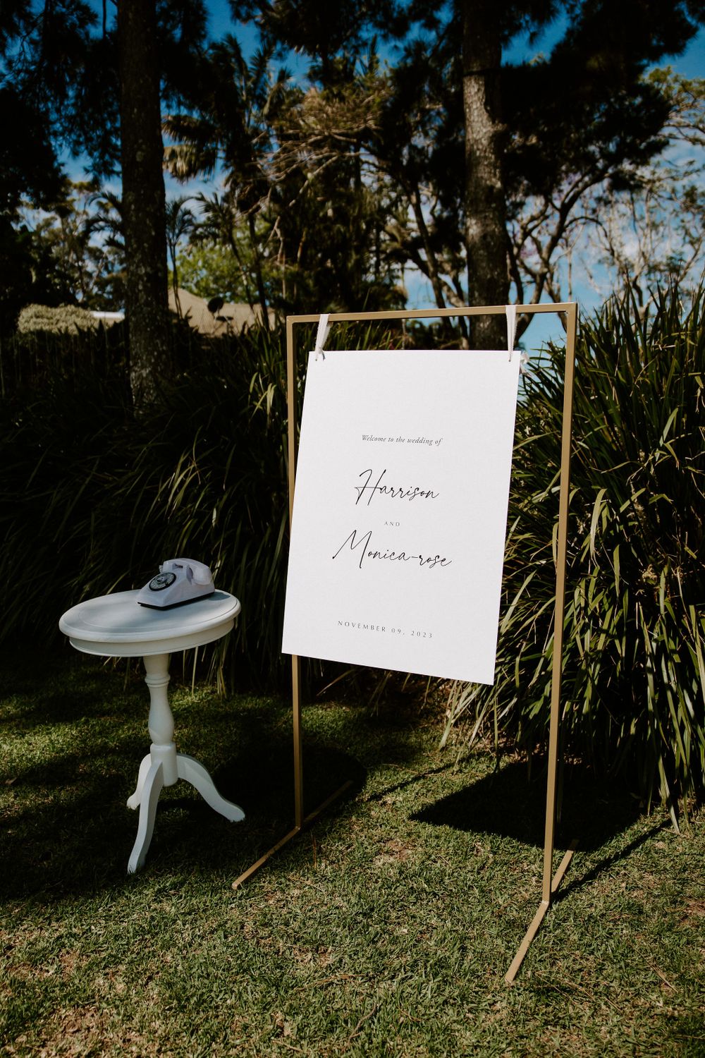 Elegant wedding welcome sign on gold stand next to white vintage side table in garden setting.