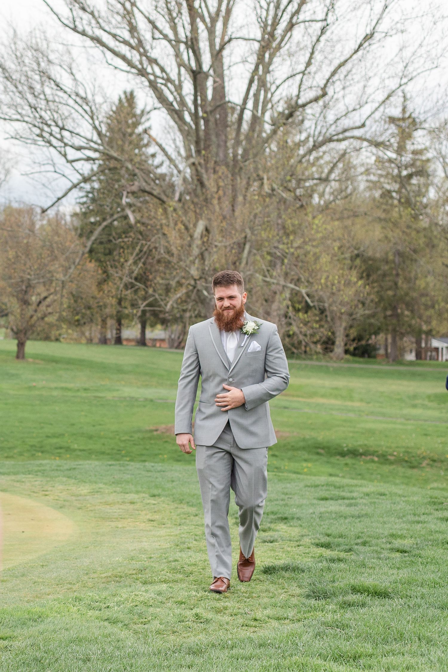 A groom in a light gray suit walks across a lush green lawn with trees in the background.
