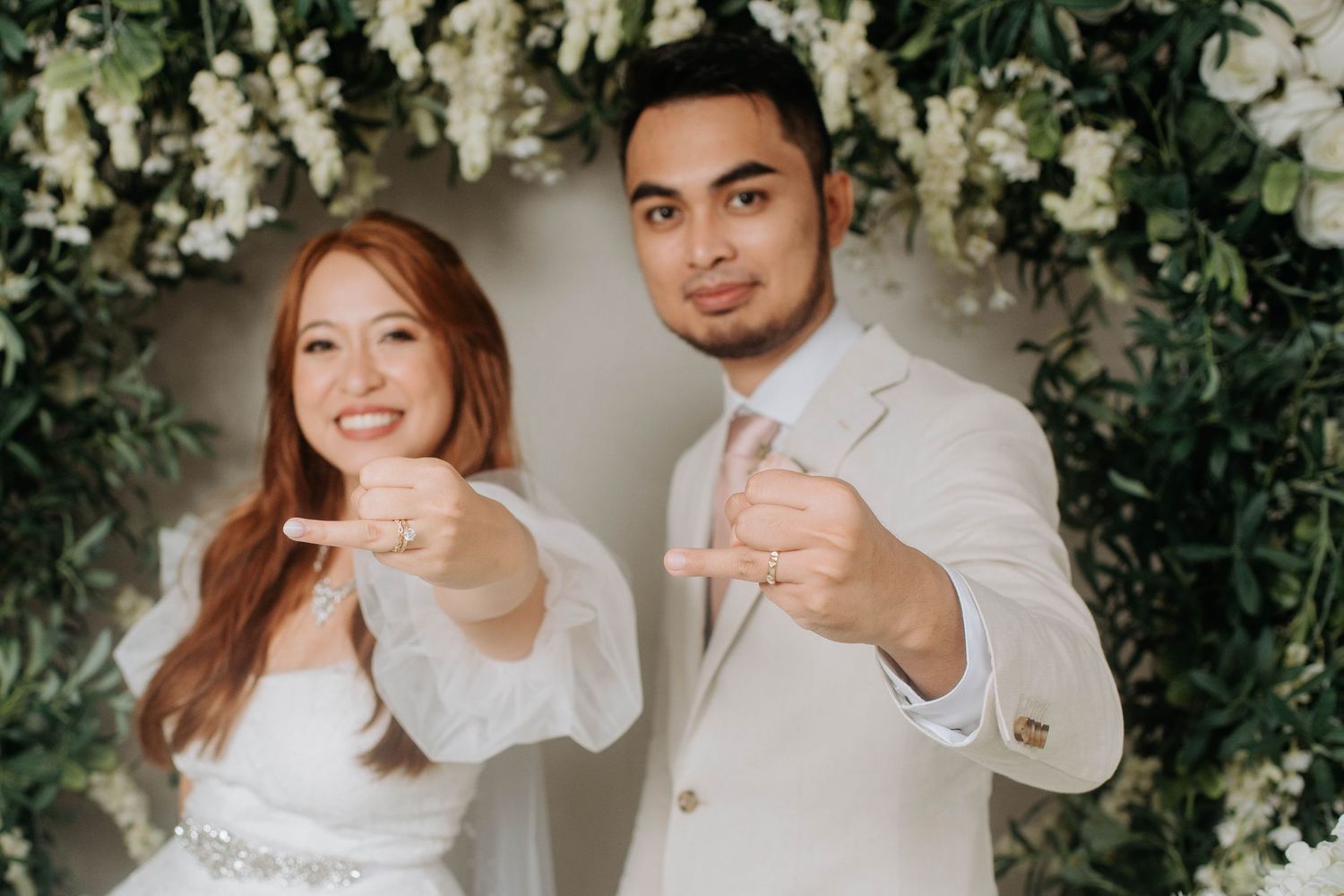 Wedding couple points playfully at camera in front of floral wall backdrop.