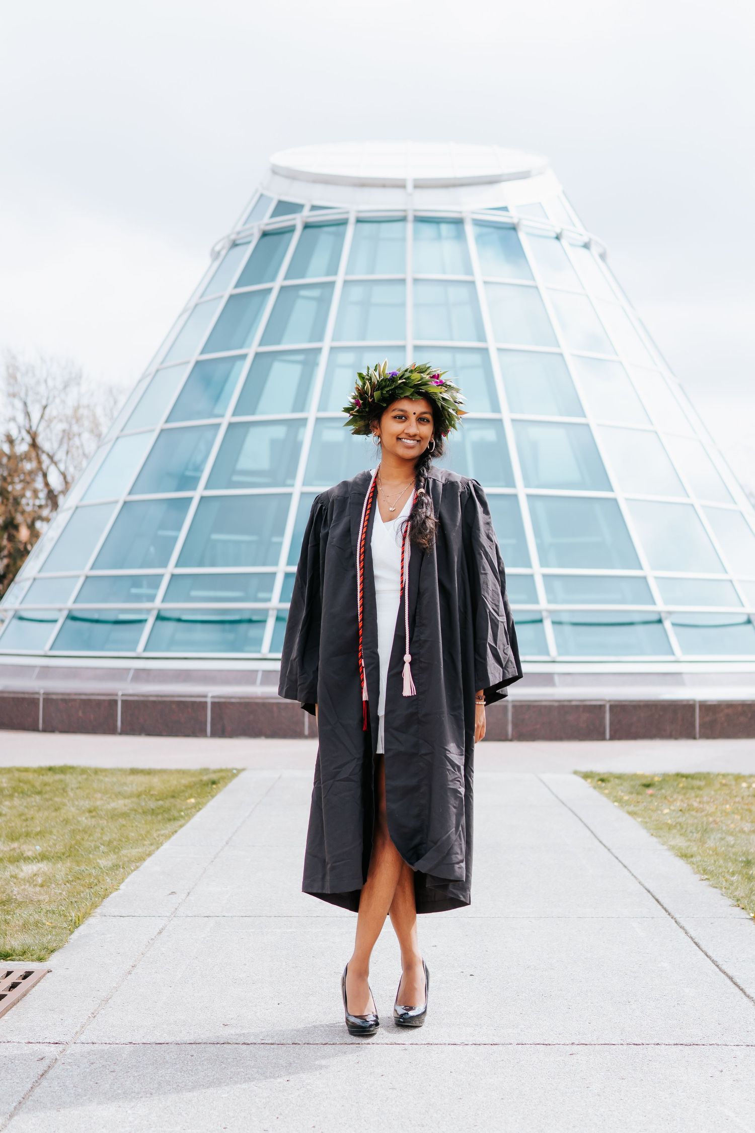Graduate in black gown and leafy crown stands in front of glass building.